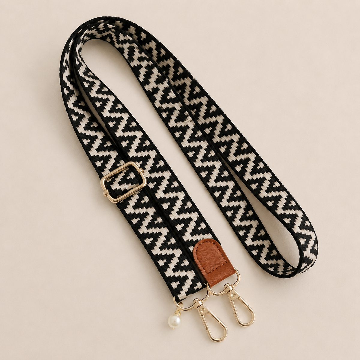 Folie à Trois Wynter long wholesale phone strap in a black and white zigzag weave with a tan leather tab, gold clasps and a pearl charm, coiled on a cream background.