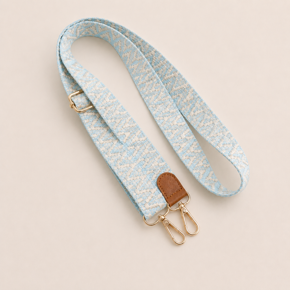 Folie à Trois Wynter long wholesale phone strap in a light blue and cream zigzag weave with a tan leather tab and gold clasps, coiled on a cream background.
