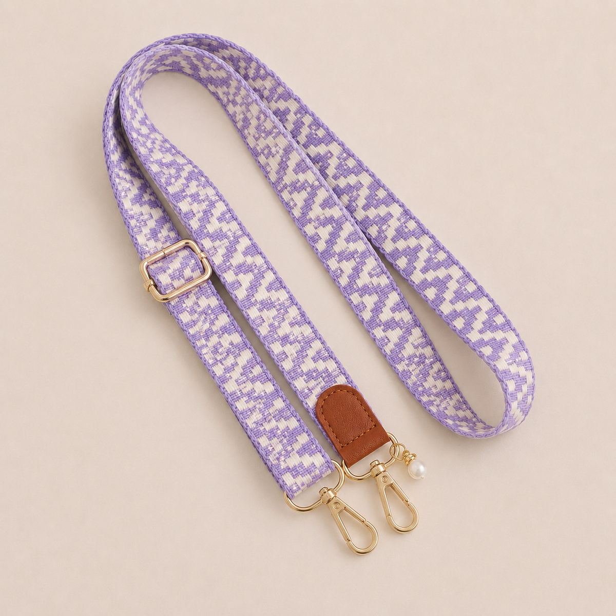 Folie à Trois Wynter long wholesale phone strap in a lilac and white zigzag weave with a tan leather tab, gold clasps and a pearl charm, coiled on a cream background.