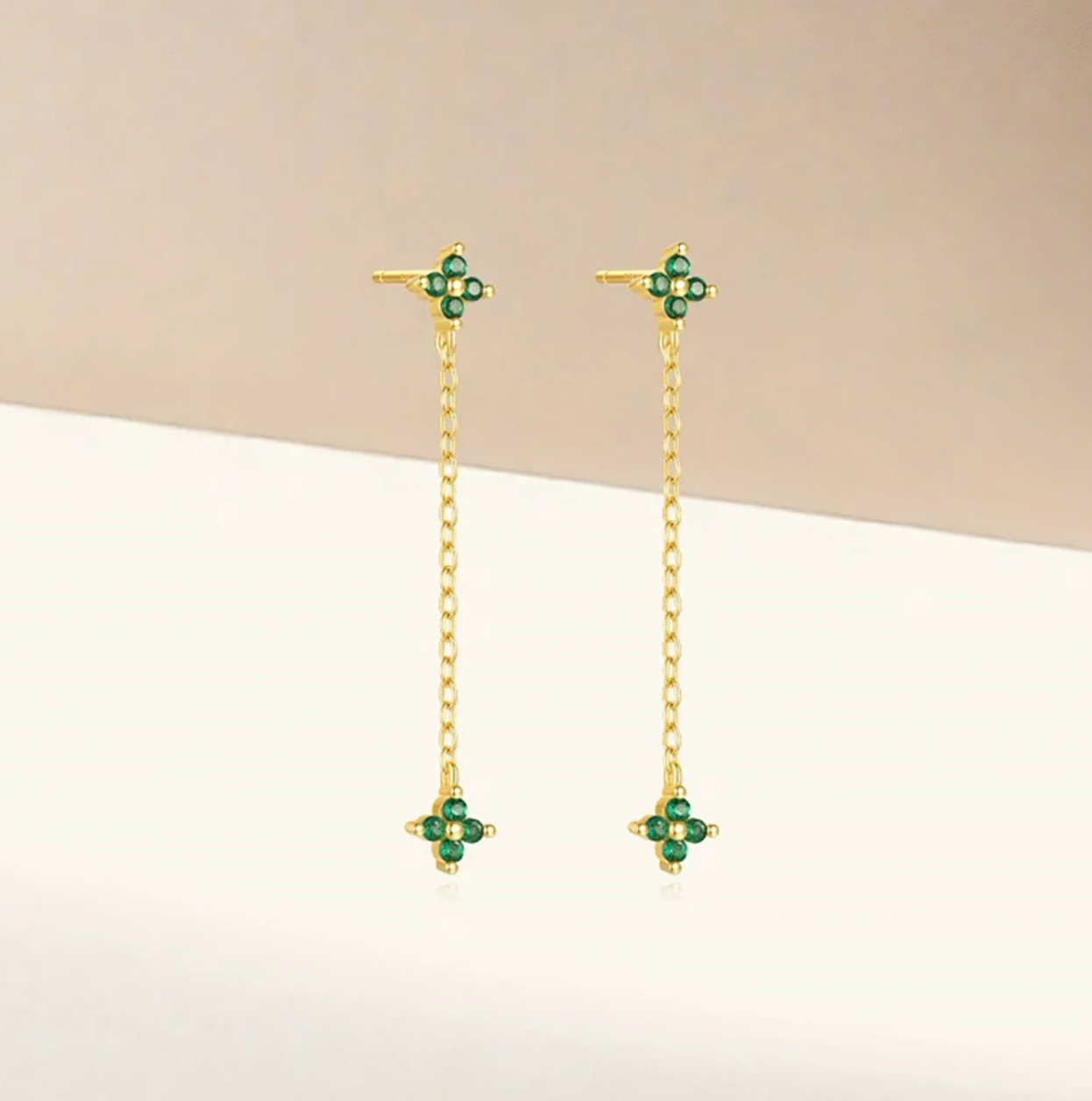 Zabbarra Earrings styled against a soft beige backdrop, the delicate chain linking two green clover crystal charms.