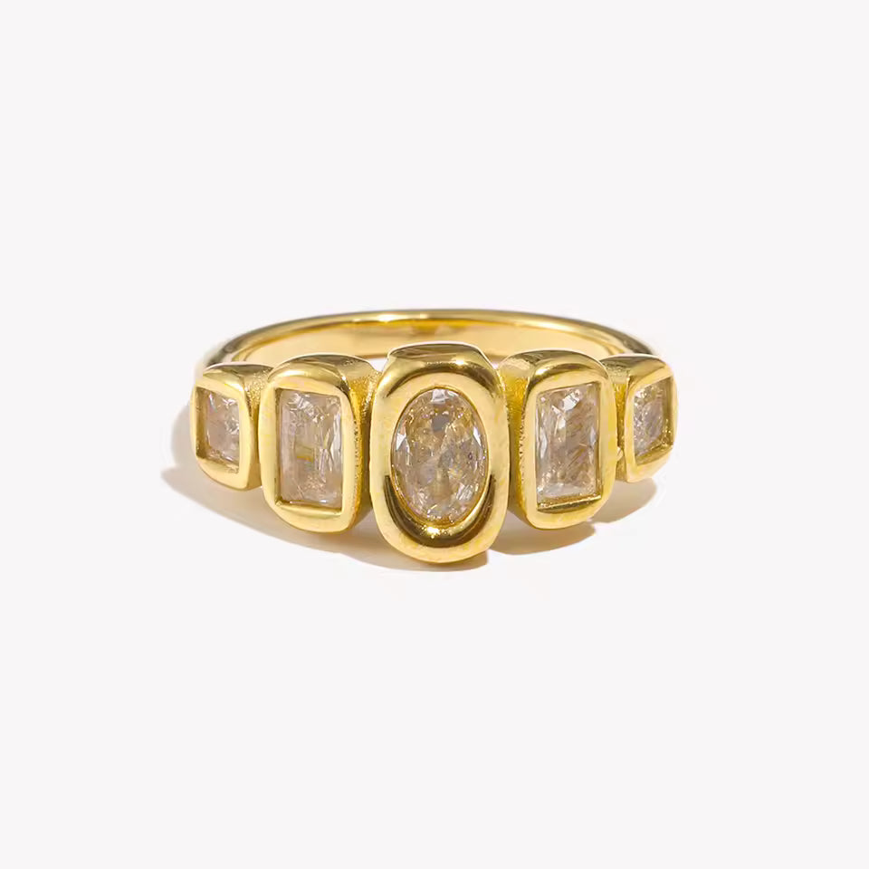Gold ring set with five bezel-set clear stones of mixed oval and rectangular cuts in a row, shown on a cream backdrop — Zippora RING.