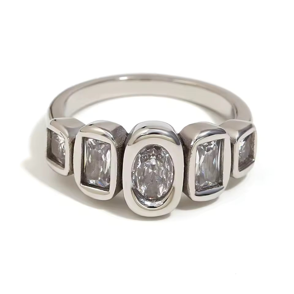 Silver ring set with five bezel-set clear stones of mixed oval and rectangular cuts in a row, shown on a white backdrop.