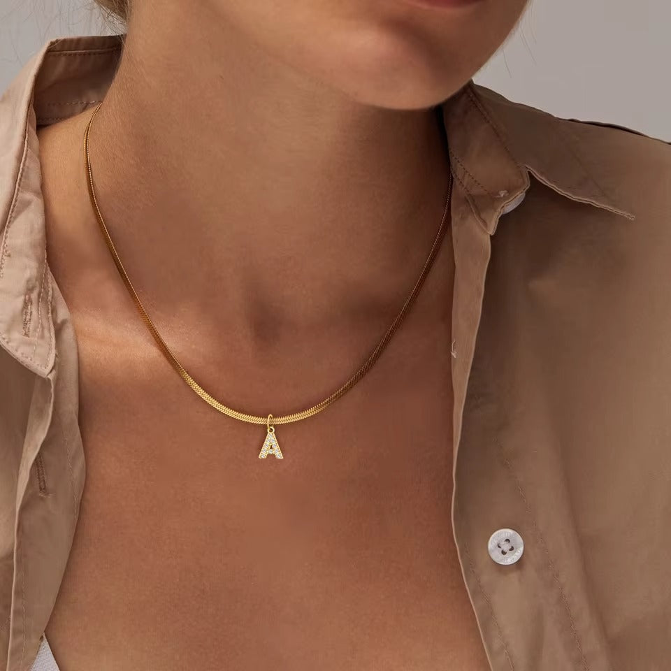 The Zircon Letter CHARM in A, a zircon-set gold initial worn at the neckline on a herringbone chain against a tan shirt.