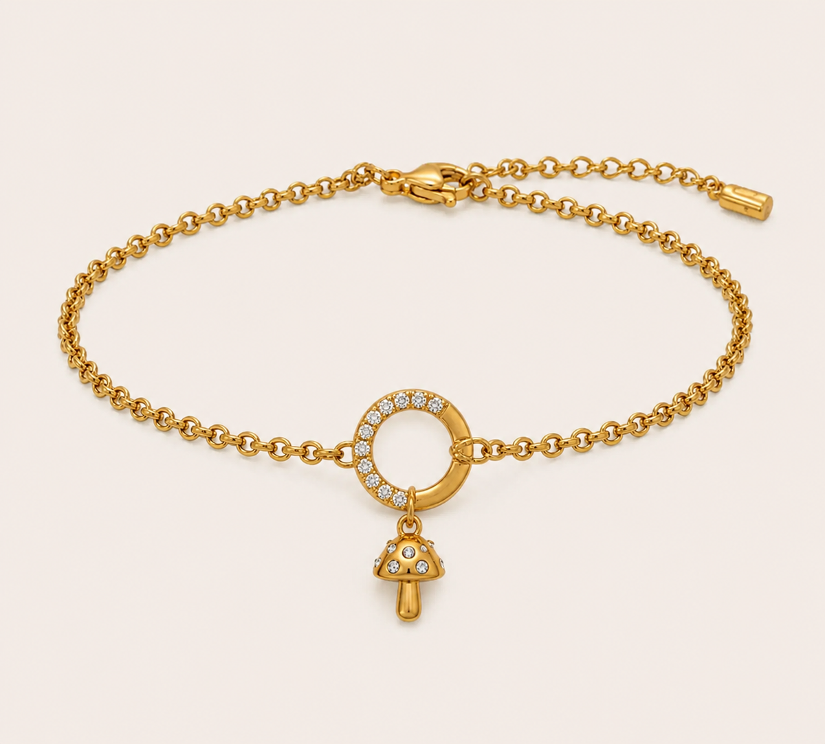 A gold chain bracelet with a circular ring set with small clear stones and a dangling mushroom-shaped charm, also accented with clear stones, shown on a light beige background.