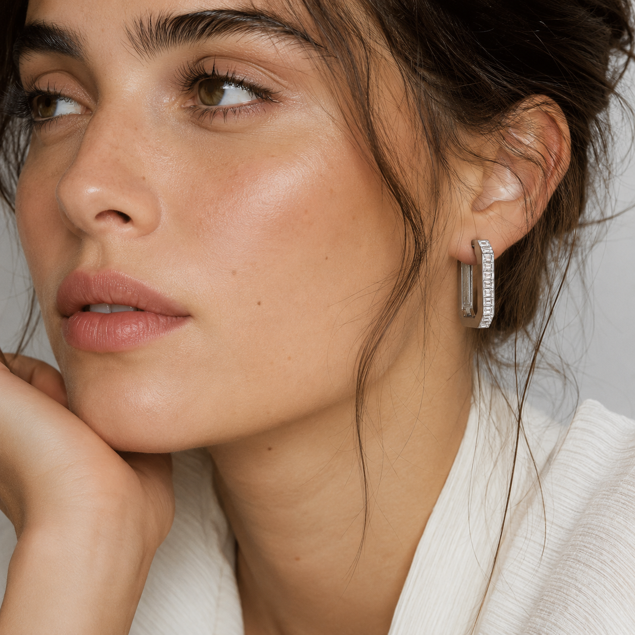 A close-up of a woman wearing a silver hoop earring adorned with small clear stones, set against a soft light gray background.