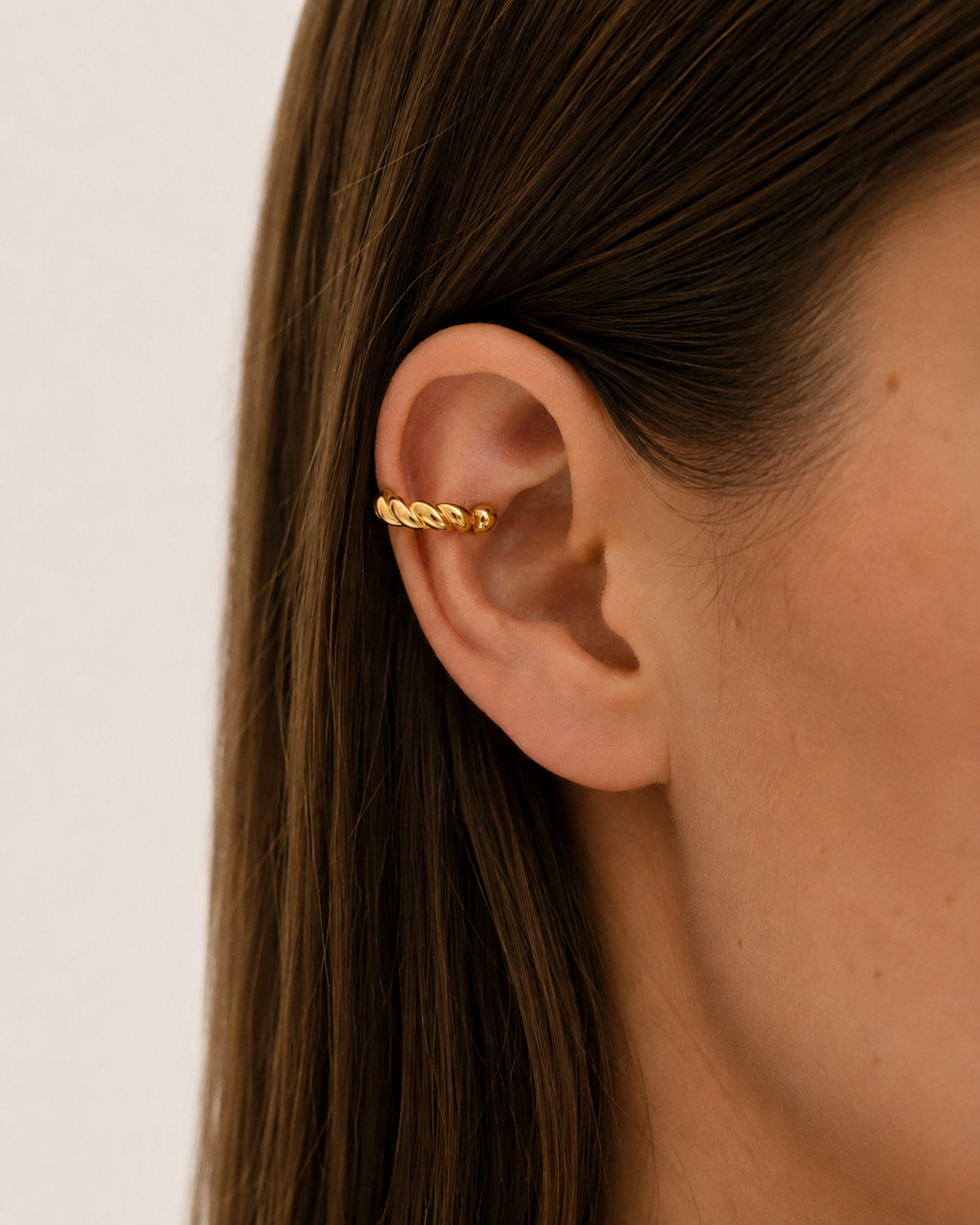 A close-up of a woman's ear wearing a delicate gold ear cuff with a twisted rope design, set against a clean, light off-white background.