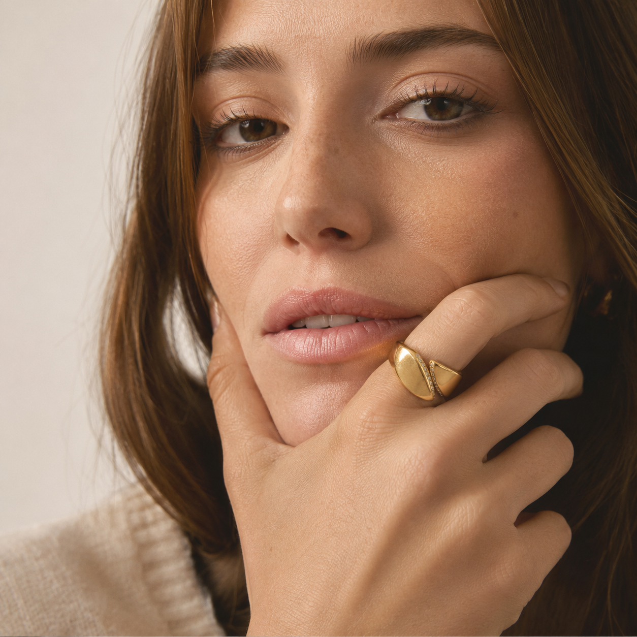 A close-up lifestyle shot of a woman wearing a chunky, polished gold ring on her finger, resting her chin on her hand against a soft beige background.