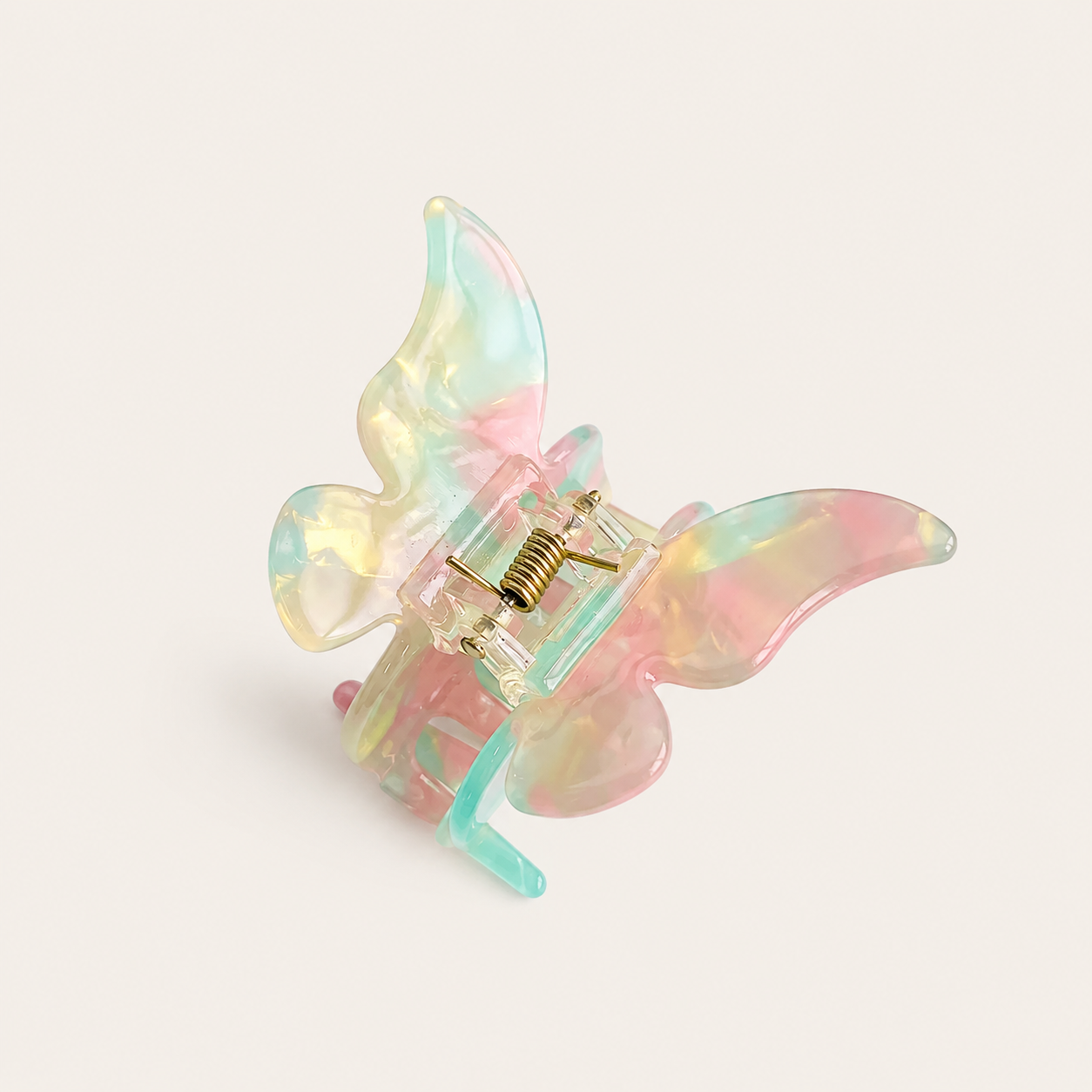 A translucent butterfly-shaped hair claw clip with pastel pink, yellow, and light blue iridescent colors, featuring a gold metal spring and clear plastic base, set against a solid off-white background.