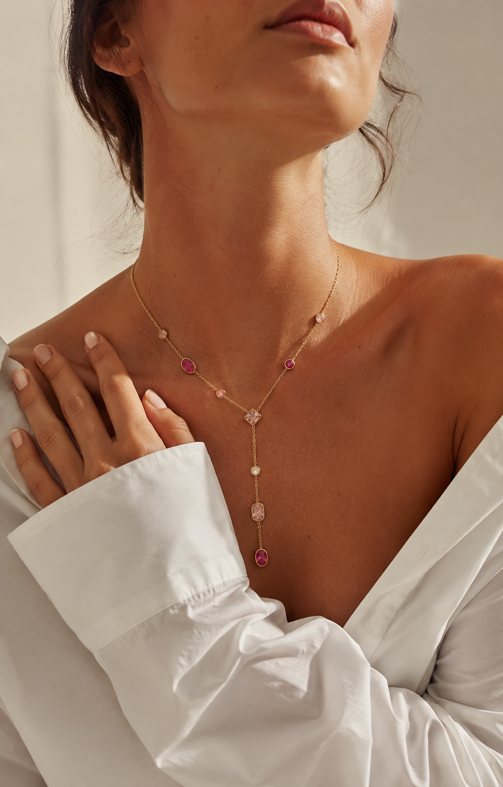 A close-up lifestyle shot of a model wearing a delicate gold Y-necklace with pink gemstone accents and clear crystal drops, paired with a white off-the-shoulder blouse.