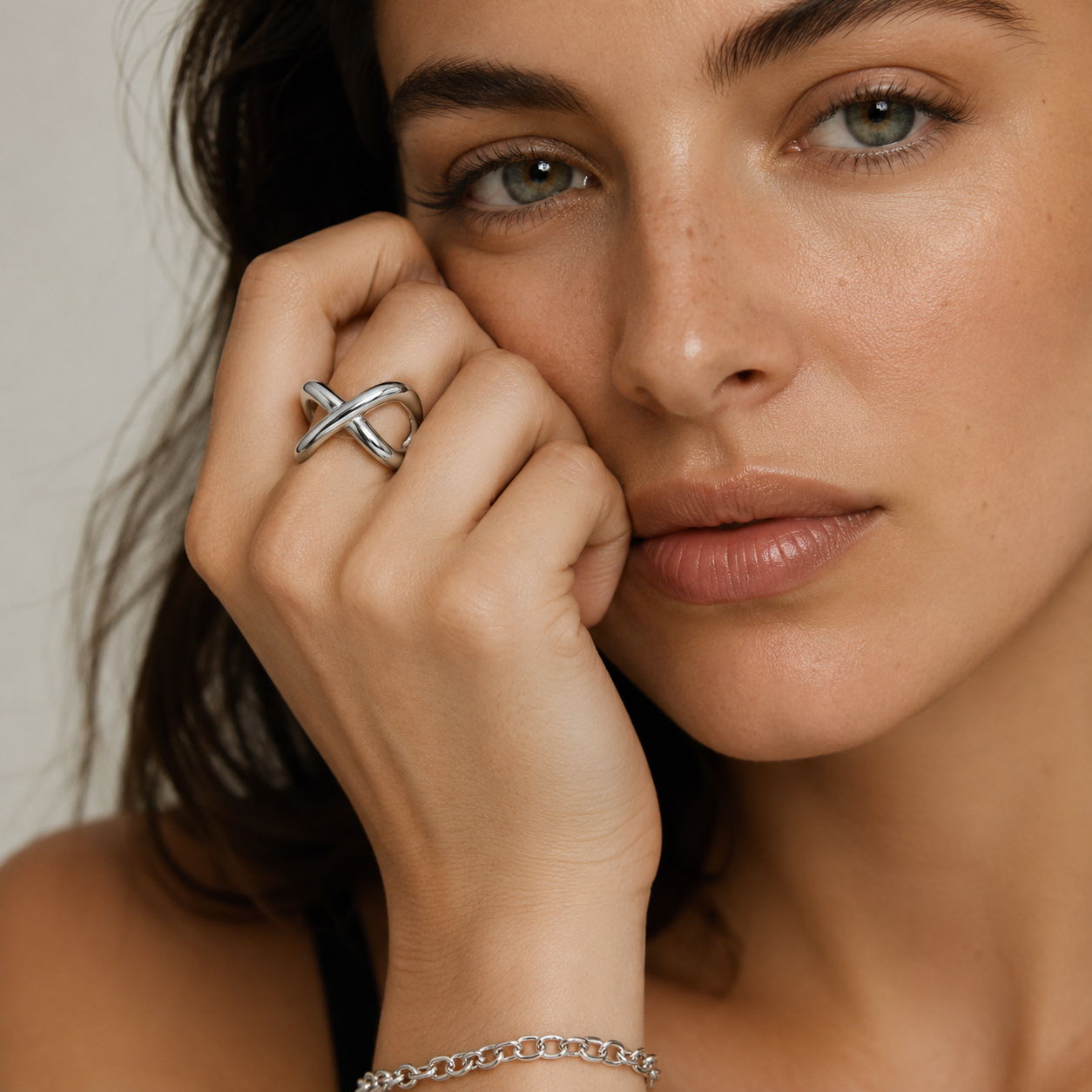 A close-up lifestyle shot of a woman wearing a silver infinity-style ring on her finger and a matching silver chain bracelet on her wrist, set against a soft, light beige background.