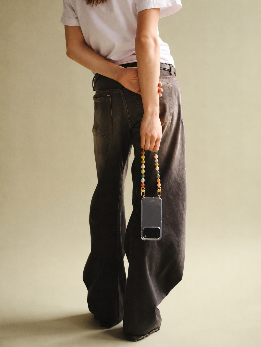 A model carries her phone via the multicolor Sable PHONE CHAIN clipped to a clear case, styled against dark denim trousers.