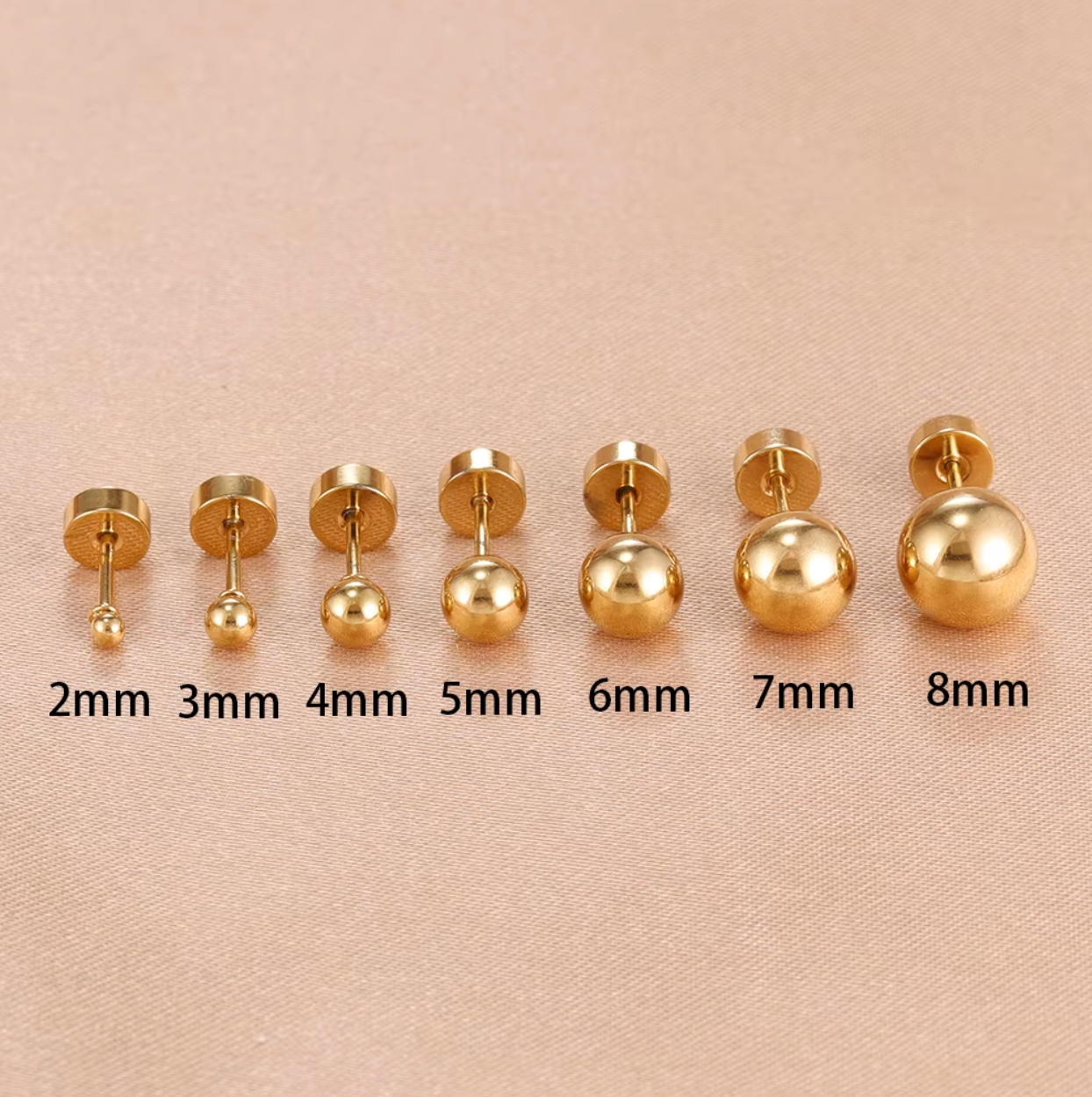 Row of gold ball stud piercings in multiple sizes displayed on a soft fabric background, showing smooth polished finish and flat screw-back closures.